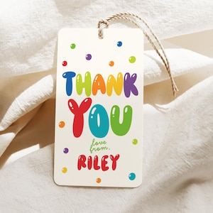 Puede incluir: Una etiqueta de regalo rectangular con las palabras "THANK YOU" en letras de burbujas coloridas. La etiqueta también incluye el texto "love from, Riley" en una fuente lúdica, con lunares coloridos. La etiqueta está unida a una cuerda natural.