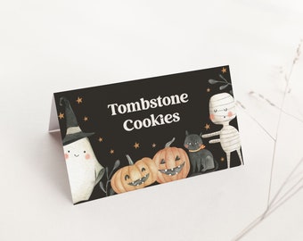 May include: A black rectangular folded card featuring the white text Tombstone Cookies. The design includes watercolor illustrations of a ghost in a hat, two jack-o'-lantern pumpkins, a black cat, and a mummy, all accented with small orange stars.