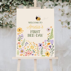 Puede incluir: Bienvenido a Amelia's First Bee-Day, un cartel con una abeja de dibujos animados y un borde floral. El cartel est&aacute; en un caballete de madera.