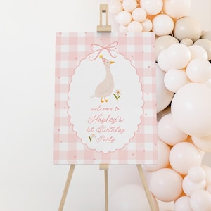 May include: A pink and white gingham sign on a wooden easel. The sign features an illustration of a goose wearing a party hat and the text "welcome to Hayley's 1st Birthday Party". A balloon arch in shades of white and beige is in the background.