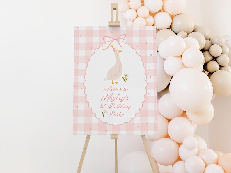 May include: A pink and white gingham sign on a wooden easel. The sign features an illustration of a goose wearing a party hat and the text "welcome to Hayley's 1st Birthday Party". A balloon arch in shades of white and beige is in the background.
