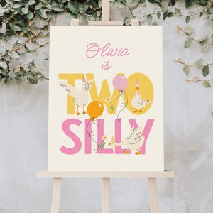 May include: A cream-colored sign on an easel with the text "Olivia is TWO SILLY." The sign features illustrations of geese wearing party hats and holding balloons. The text is in pink and yellow.