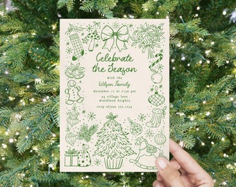 May include: A cream-colored Christmas invitation with green illustrations and text. The invitation features a Christmas tree, ornaments, and the text "Celebrate the Season with the Wilson Family". The invitation is held in front of a Christmas tree.