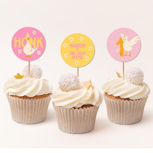 May include: Three cupcakes with white frosting, each topped with a decorative pick. The picks feature a goose illustration and text that reads "Honk", "Harper is one silly goose", and "1". The cupcakes have a white and gold star and a white ball on top.