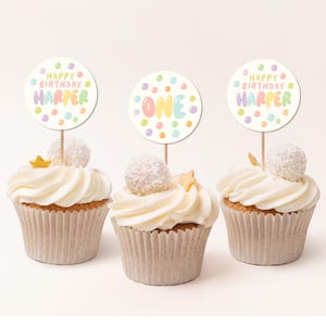 May include: Three cupcakes with white frosting and colorful sprinkles. Each cupcake has a round, white paper topper with colorful polka dots and text that reads "Happy Birthday Harper", "One", and "Happy Birthday Harper".