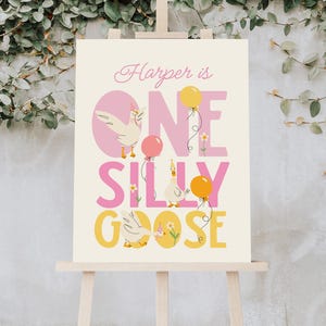 May include: A cream-colored sign on an easel with the text "Harper is ONE SILLY GOOSE." The design includes illustrations of geese, balloons, and flowers in pink, yellow, and cream colors. The sign is displayed against a backdrop of green foliage.