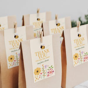 Puede incluir: Bolsas de regalo de papel marr&oacute;n con etiquetas de agradecimiento amarillas y blancas con una abeja de dibujos animados y dise&ntilde;os florales. Las etiquetas dicen "&iexcl;Gracias por estar aqu&iacute;!" y "&iexcl;Gracias por estar aqu&iacute;! Con cari&ntilde;o, Amelia".