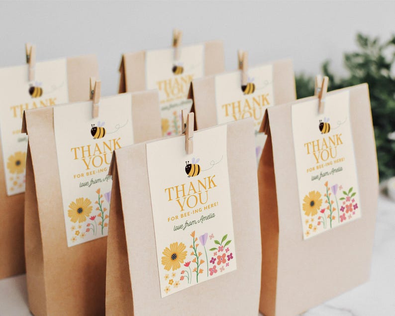 Puede incluir: Bolsas de regalo de papel marr&oacute;n con etiquetas de agradecimiento amarillas y blancas con una abeja de dibujos animados y dise&ntilde;os florales. Las etiquetas dicen "&iexcl;Gracias por estar aqu&iacute;!" y "&iexcl;Gracias por estar aqu&iacute;! Con cari&ntilde;o, Amelia".