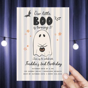 May include: A rectangular birthday invitation card with a light gray and white vertical striped background. It features a central ghost illustration holding a small pumpkin bucket, surrounded by bats and orange stars, with the text Our little BOO is turning 2! Join us to celebrate Freddie's 2nd Birthday, October 28th at 3 PM, 123 Cherry Drive, Woodland Heights, RSVP by October 24th, Jenny 555.5555.