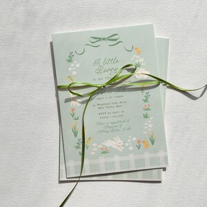 Pu&ograve; includere: Un invito per una festa di nascita di colore azzurro chiaro con un nastro verde legato intorno. L'invito presenta un motivo floreale con un coniglietto e il testo "A little bunny is on the way".