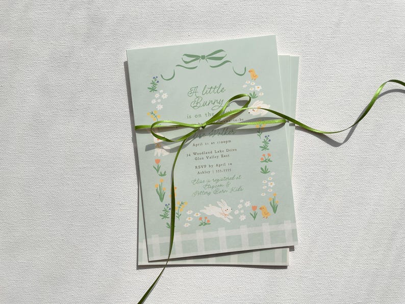 Pu&ograve; includere: Un invito per una festa di nascita di colore azzurro chiaro con un nastro verde legato intorno. L'invito presenta un motivo floreale con un coniglietto e il testo "A little bunny is on the way".
