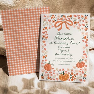 Può includere: Un biglietto d'invito con un design a ghirlanda floreale e il testo "Our little Pumpkin is turning One!". La carta presenta zucche arancioni e un fiocco. La busta ha un motivo a quadretti pesca e bianco.