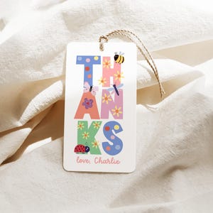 May include: A white gift tag with the word "THANKS" in colorful, whimsical letters. The letters are decorated with flowers, butterflies, a ladybug, and a bee. The tag includes the text "love, Charlie" at the bottom.