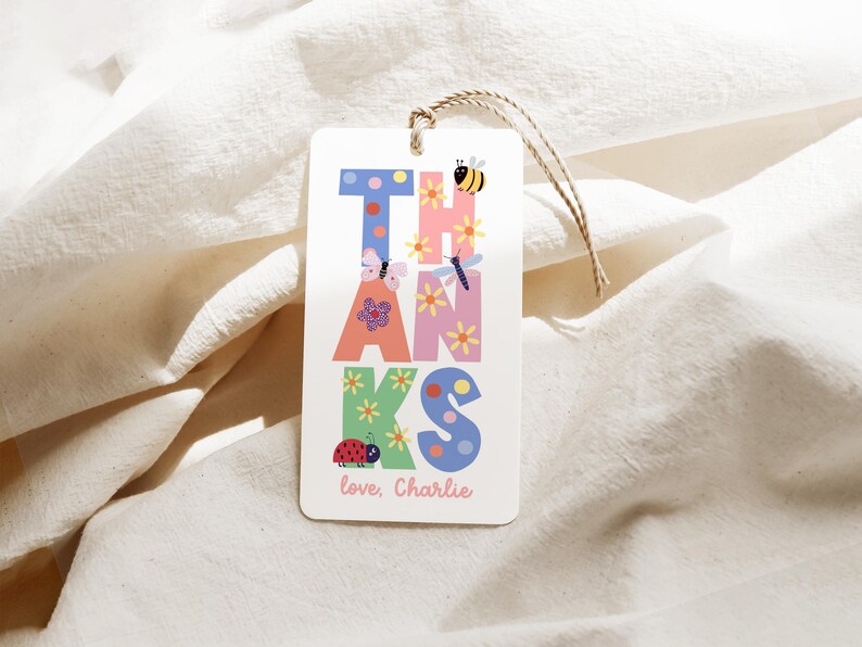 May include: A white gift tag with the word "THANKS" in colorful, whimsical letters. The letters are decorated with flowers, butterflies, a ladybug, and a bee. The tag includes the text "love, Charlie" at the bottom.