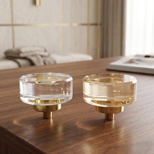 Puede incluir: Dos pomos circulares para armarios con tapas de cristal transparente y ámbar sobre bases de metal dorado. Cada pomo tiene un cuerpo cilíndrico de cristal con una superficie superior plana y una hendidura circular central.