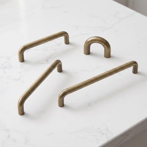May include: A collection of four cabinet hardware pieces in a brushed antique brass finish. The set includes three elongated bar pulls of varying lengths and one small, arched U-shaped knob, all featuring a smooth, rounded cylindrical profile.