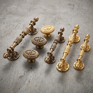 May include: A collection of brass-toned metal hardware including three elongated cabinet pulls with decorative floral-inspired detailing and three oval-shaped knobs featuring a central star-like motif surrounded by a textured border.