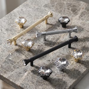 May include: A collection of cabinet hardware including three long, fluted metal drawer pulls in gold, silver, and black finishes, paired with five matching flower-shaped knobs featuring clear, faceted crystal centers set in metal bases.