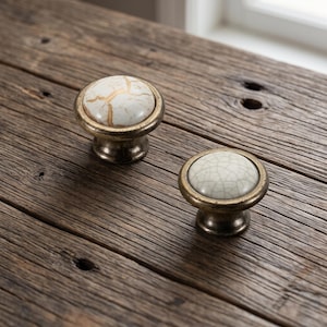 May include: Two round cabinet knobs featuring a metallic bronze-toned base and a smooth, domed top. One knob displays a white surface with prominent gold-colored veining, while the other features a light grey surface with a fine crackle pattern.