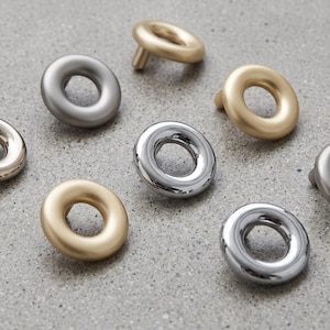 Puede incluir: Una colección de pomos circulares para armarios en varios acabados, incluyendo oro mate, plata cepillada y cromo pulido. Cada pomo presenta una forma de anillo suave y redondeada con una abertura central y un pequeño poste de montaje.