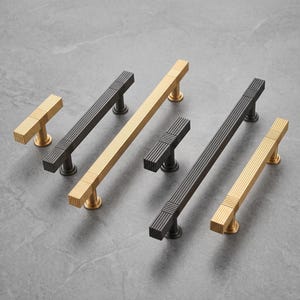May include: A collection of six cabinet pulls in matte black and brushed gold finishes. Each handle features a linear ribbed texture along the bar and a square-shaped end cap. The hardware includes both T-bar style knobs and longer drawer pulls.