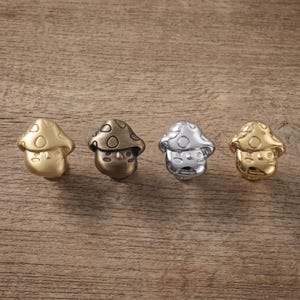 May include: Four mushroom-shaped cabinet knobs arranged in a row. Each knob features a rounded cap with circular indentations and a face with simple eyes and a mouth. The finishes include matte gold, antique bronze, polished silver, and shiny gold.