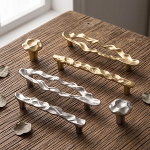 May include: A collection of cabinet hardware including two gold-toned and two silver-toned wavy drawer pulls, along with one gold-toned and one silver-toned mushroom-shaped knob, all featuring a textured, organic, and uneven metallic surface finish.