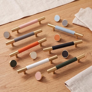Pode incluir: Uma coleção de ferragens para armários com puxadores de barra e puxadores circulares em metal dourado. Os puxadores apresentam inserções de várias cores, incluindo rosa, cinzento, laranja, preto, branco e verde escuro, com acabamentos lisos e texturizados.