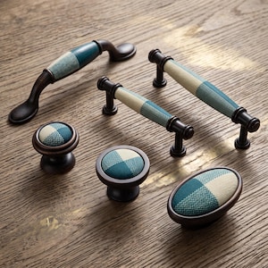 May include: A collection of dark bronze cabinet hardware including two arched handles, two cylindrical bar pulls, two round knobs, and one oval knob. Each piece features a central inlay of textured fabric in a checkered pattern of teal and cream.