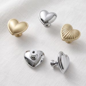 May include: A collection of five heart-shaped cabinet knobs in various finishes. Included are a matte gold smooth heart, a polished silver smooth heart, a ribbed gold heart, a polished silver heart shown from the back, and a side view of a silver knob.