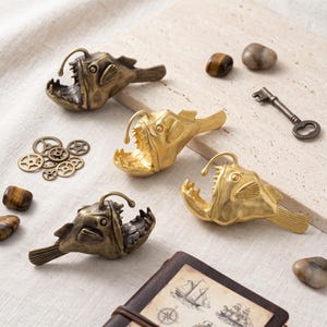May include: Four metallic anglerfish figurines with open mouths and prominent teeth. Two are finished in a dark antique bronze tone, and two are in a bright polished gold tone. Each fish features a dorsal lure and detailed fin textures.