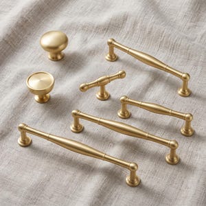 May include: A collection of brushed gold cabinet hardware including two round knobs and four bar pulls of varying lengths. Each piece features a smooth, matte metallic finish with a cylindrical design, showcasing a consistent aesthetic across all items.