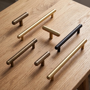 May include: A collection of seven metal cabinet pulls and knobs in various finishes including brushed gold, matte black, and antique brass. The hardware features a cylindrical bar design with rounded ends and two mounting posts for attachment.