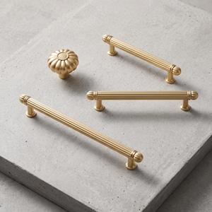May include: A set of gold-toned cabinet hardware including one round, fluted knob with a textured center and three elongated, cylindrical drawer pulls featuring vertical ribbed detailing and rounded end caps with a smooth, matte metallic finish.