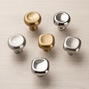 May include: A collection of six cabinet knobs arranged in a circular pattern. The set includes three round knobs and three square knobs with rounded edges, featuring brushed brass, polished chrome, and brushed nickel metallic finishes.
