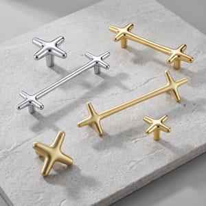 May include: A collection of cabinet hardware featuring cross-shaped knobs and elongated bar pulls. The set includes polished silver-tone metal pieces and matte gold-tone metal pieces, each with a distinct four-pointed star or cross design.