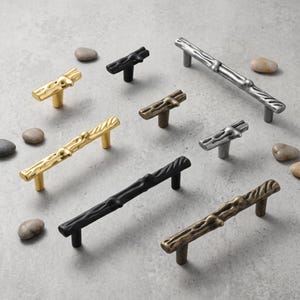 May include: A collection of cabinet hardware including T-shaped knobs and long drawer pulls with a textured, branch-like design. The hardware comes in various metallic finishes including polished gold, matte black, antique bronze, and brushed silver.