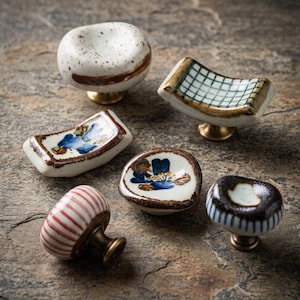 May include: A collection of six ceramic cabinet knobs featuring various designs including speckled white, blue floral patterns, a green grid, and red or blue stripes, each mounted on a brass-toned metal base with a rounded or curved shape.
