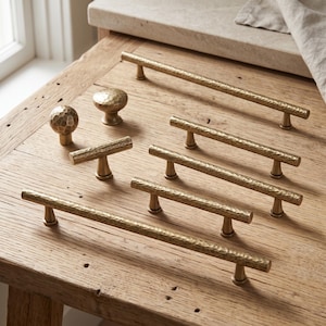 May include: A collection of brass cabinet hardware featuring a hammered texture. The set includes one long bar pull, three shorter bar pulls, one T-bar knob, and one round mushroom-shaped knob, all displaying a consistent metallic gold finish.