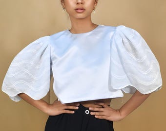 May include: A light blue cropped top with a round neckline and white puff sleeves with a wavy pattern. The top is worn with black trousers.