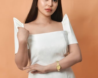 May include: A woman wearing a white dress with puffed sleeves and a gold watch. The dress has a square neckline and short sleeves. The woman is standing in front of a peach-colored background.
