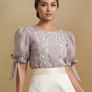 May include: A lilac-colored blouse with white embroidered detailing on the bodice and puffed sleeves tied with bows. The blouse is paired with a cream-colored skirt, creating a soft, elegant look. The outfit is suitable for formal occasions.