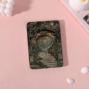 Pode incluir: Cartão retangular com uma ilustração de uma porta de hobbit. A porta verde está emoldurada por um arco de tijolo, com uma escada de pedra que leva até ela. A cena é cercada por folhagens e vinhas, criando uma estética fantasiosa.