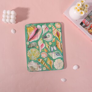 May include: A rectangular, teal-colored tray featuring a vibrant assortment of seashells in various shapes and colors, including pink, yellow, and white. The tray is adorned with a collection of seashells, creating a coastal theme.