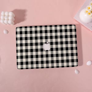 May include: A laptop case with a black and white gingham pattern. The case features the Apple logo in the center. The background is a solid pink color, with a few decorative items.