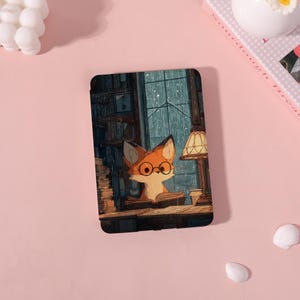 Puede incluir: Una funda negra para tableta con un zorro de dibujos animados con gafas, leyendo un libro junto a una lámpara. La ilustración tiene una paleta de colores cálidos con una biblioteca y una ventana lluviosa en el fondo.
