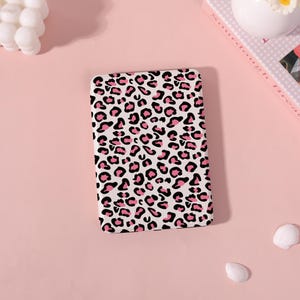 May include: A rectangular white case with a pink and black leopard print pattern. The case is on a pink surface, with a book and decorative items in the background.
