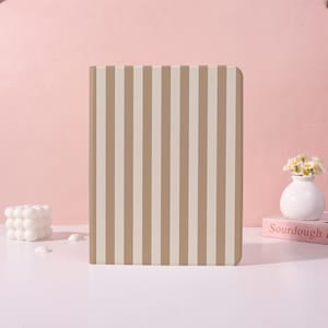 Op de afbeelding: Een rechthoekig notitieboek met een verticaal gestreept patroon in beige en gebroken wit. Het notitieboek staat rechtop op een wit oppervlak, met een roze achtergrond. Een kleine witte vaas met bloemen en een boek staan op de achtergrond.