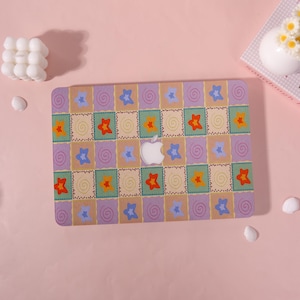 May include: A laptop case with a colorful patchwork design featuring stars and swirls in a grid pattern. The case has a white Apple logo in the center and is set against a pink background.