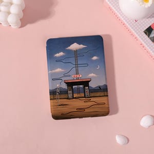 May include: A rectangular e-reader case featuring a surreal illustration of a skeleton standing in a desert landscape before a gate labeled EXIT. A long ladder extends from the gate toward a single cloud in a blue sky, with winding lines across the scene.